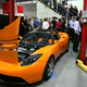 A photo of an orange Tesla Roadster on display in a crowd.